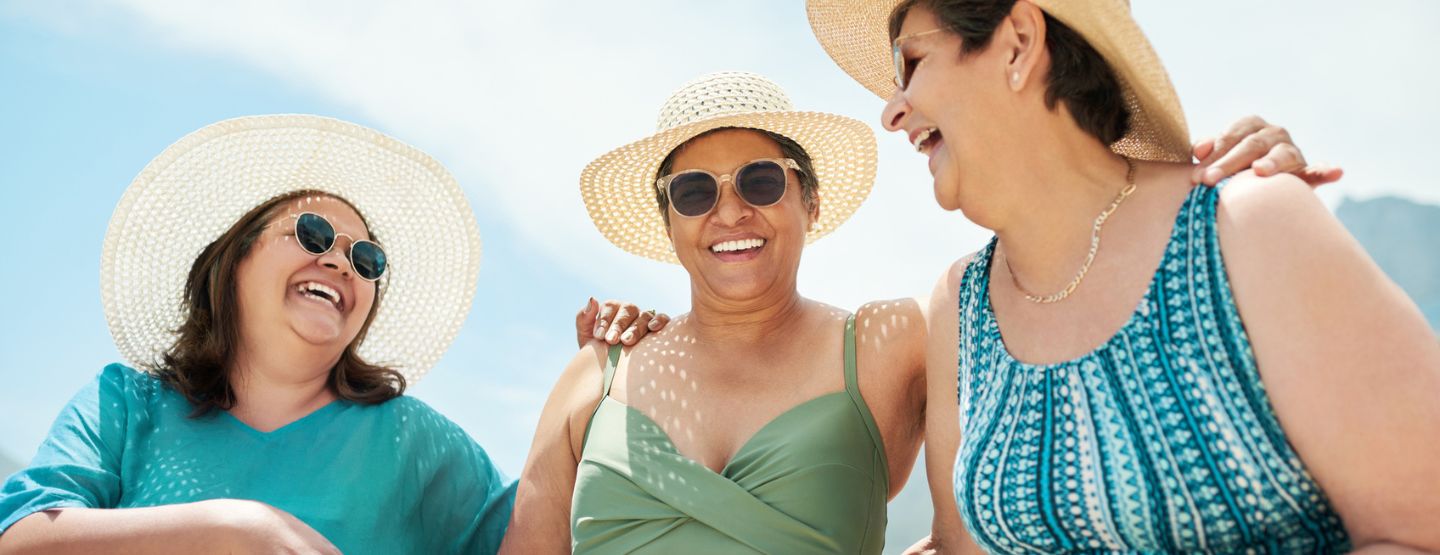 Three mature women friends smiling at each other wearing swimwear, sunglasses and wide-brimmed hats.