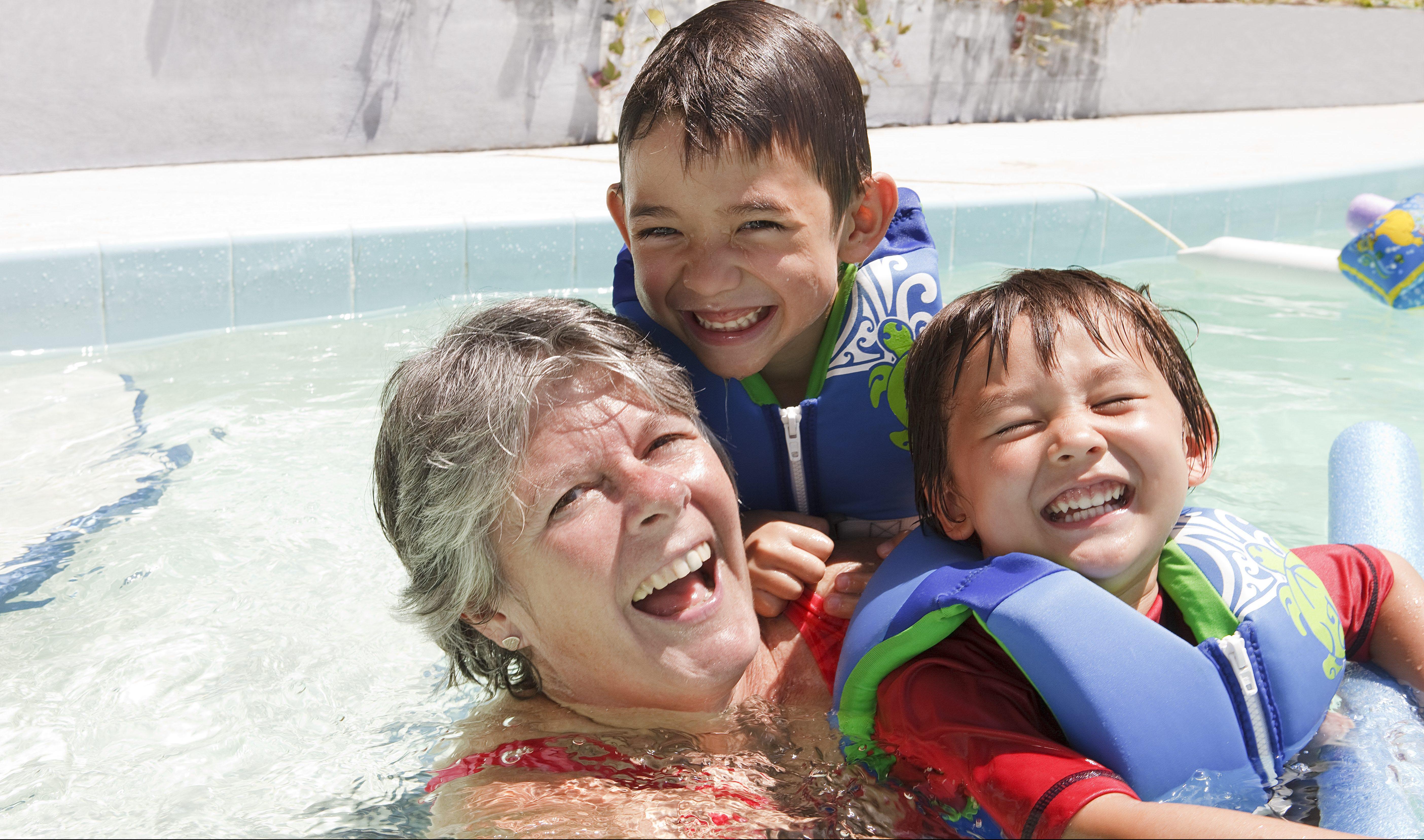 A grandmother swimming in a pool while holding her two grandchildren.