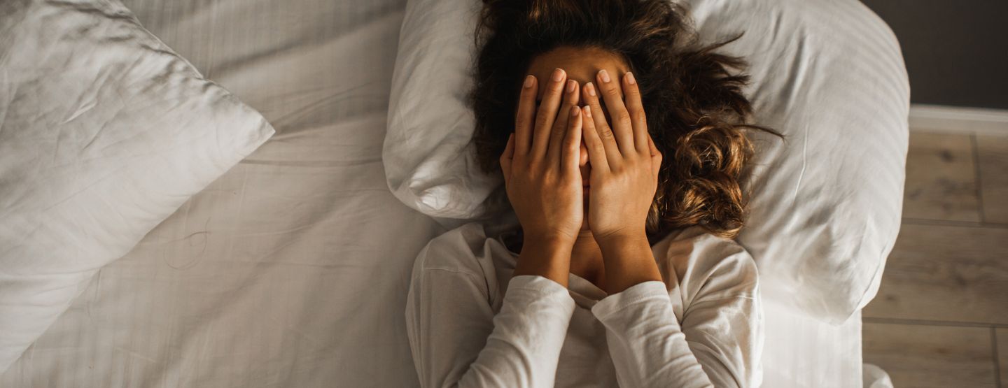 Woman laying on back in bed with hands over eyes. 