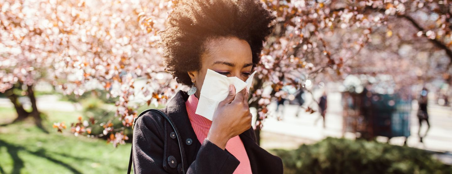 Woman walking outside under blooming tree while blowing nose. 