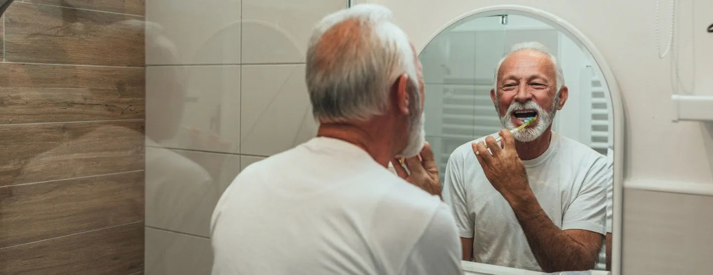 Mature man smiling at mirror while brushing teeth.