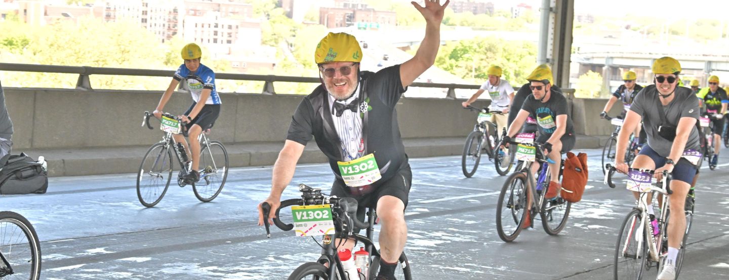 Bradley waving as he rides by on a bicycle during a race. 