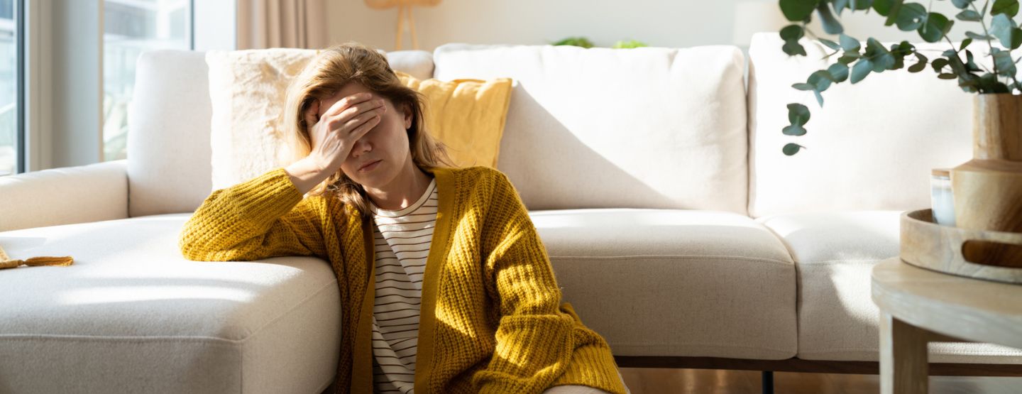 Woman sitting on the ground by couch holding head in discomfort. 