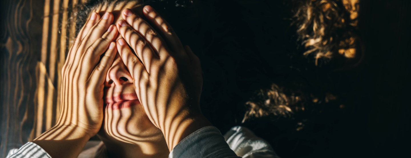 Woman covers eyes in discomfort as light shines through the blinds on her face. 
