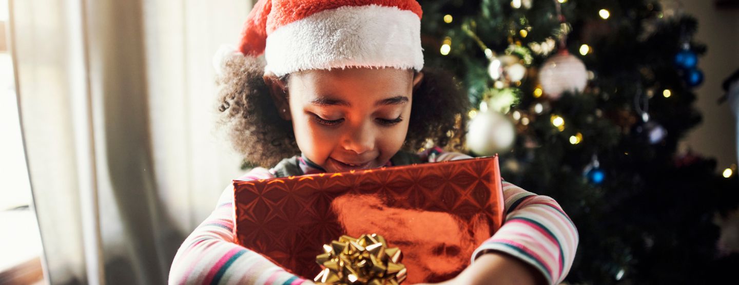 Young girl holding wrapped gift in front of Christmas tree.