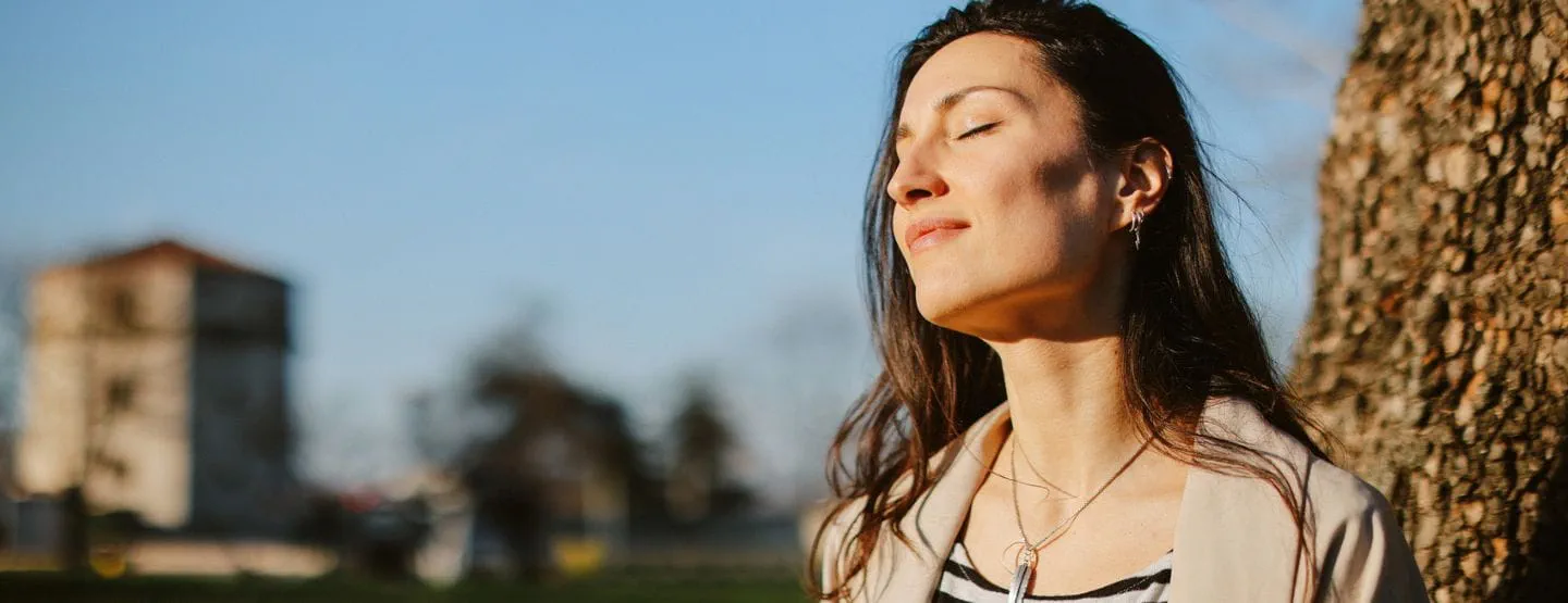 Woman sitting against tree smiling with eyes closed into the sun.