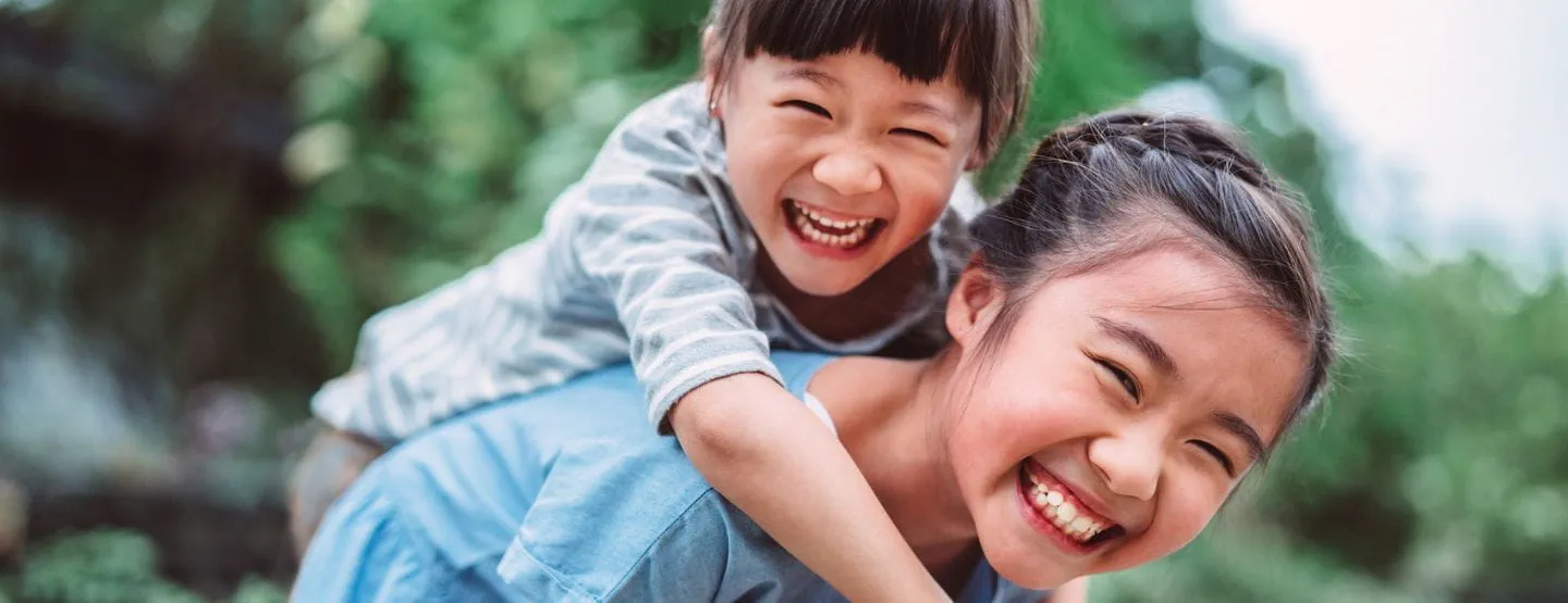 Older sister giving younger sister a piggy back ride while smiling at the camera. 