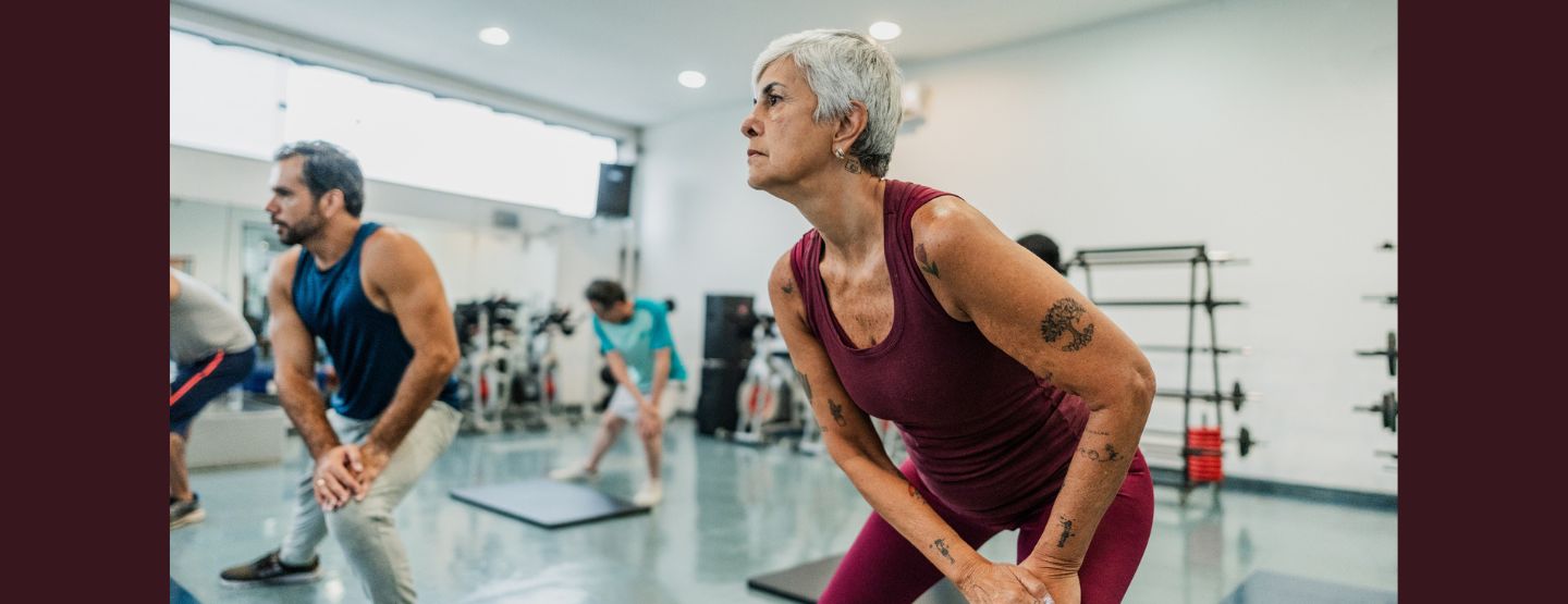 Older woman with short grey hair doing a standing stretch in a class with others stretching.