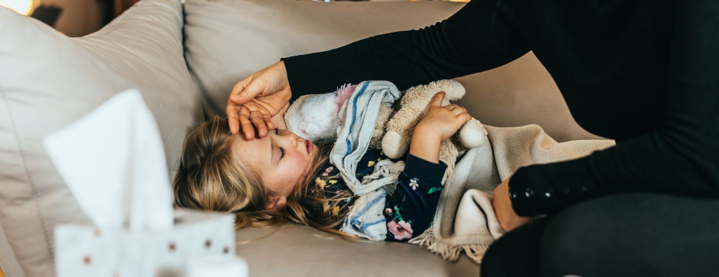 Young child lays ill on couch while mother feels her head for a fever.