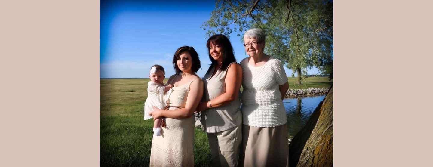 Four generations pose in front of a pond and field of grass. Alyson Stanton holds her baby daughter. To the right is her mother, Cindy Lau, and her grandmother, Jan Anaple. All three women are wearing beige outfits and smiling at the camera.