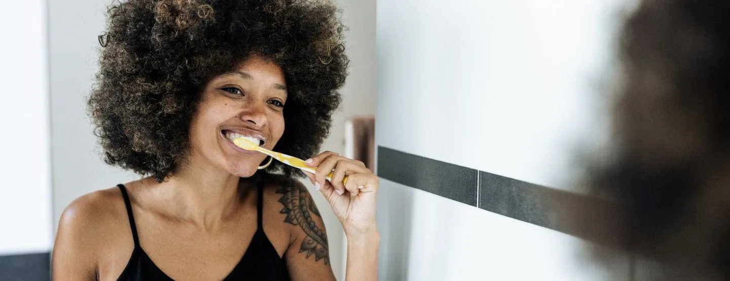 Woman standing in bathroom brushing teeth while looking in mirror.