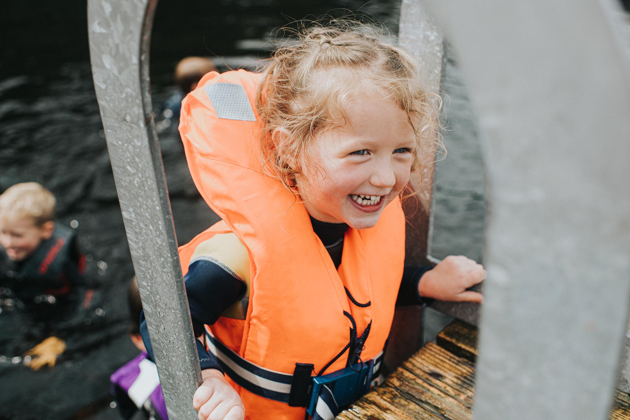 Happy and confident pre-school age girl climbing up a ladder onto a jetty wearing a fluorescent orange life jacket.