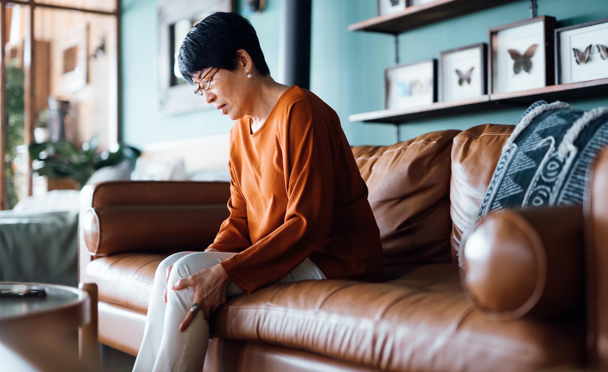 A distraught senior Asian woman feeling unwell, suffering from pain in leg while sitting on sofa in the living room at home