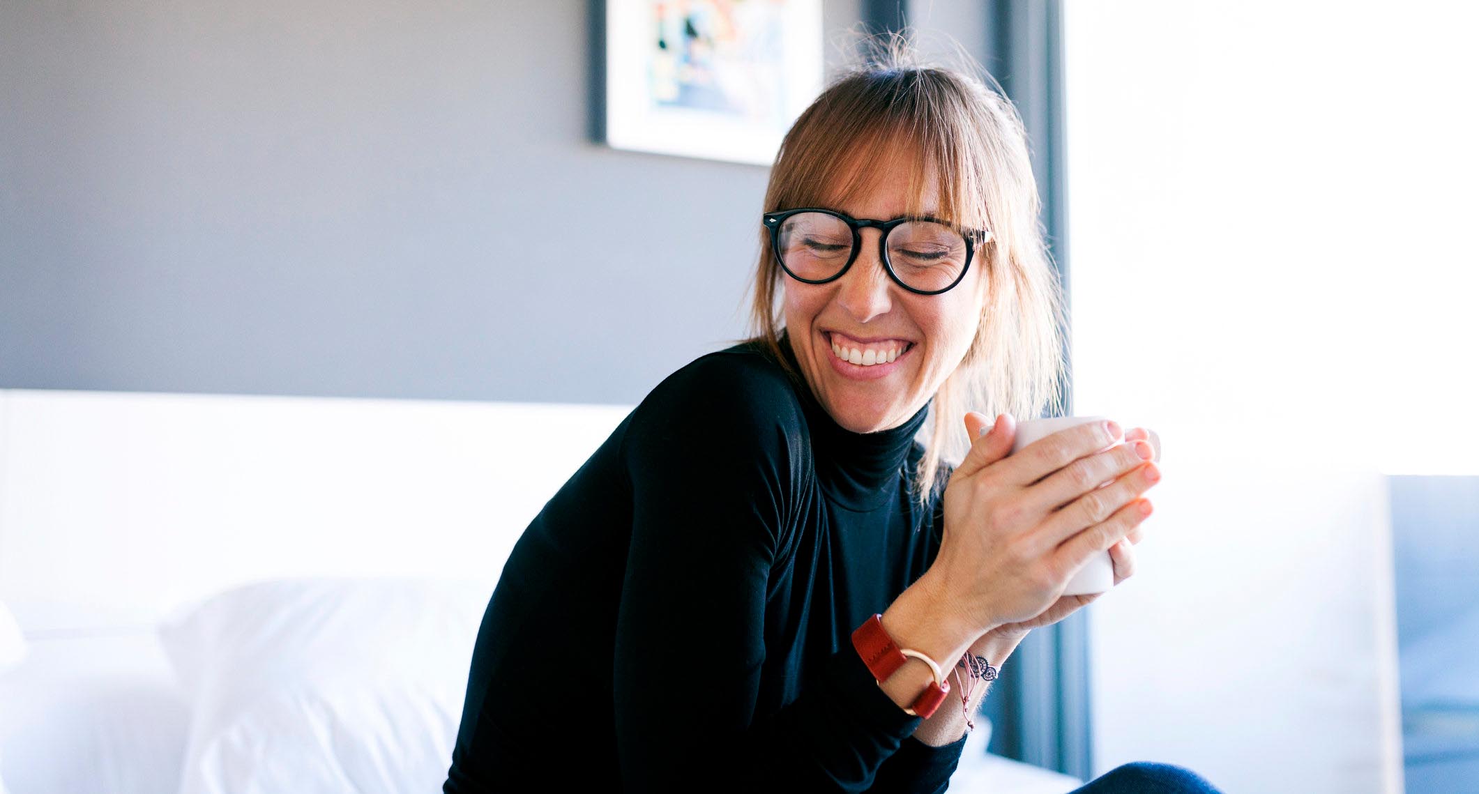 A woman with glasses smiles while sitting on her bed and holding a mug in her hands.