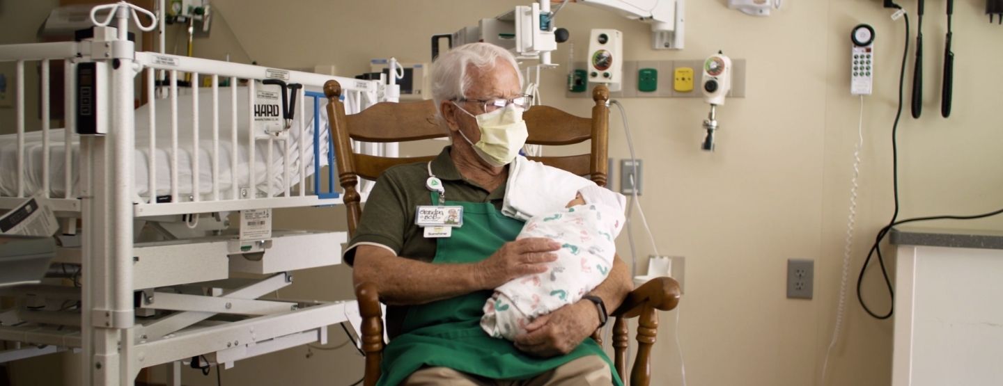 Grandpa Bob Easler sits in a wooden rocking chair, holding a swaddled infant.