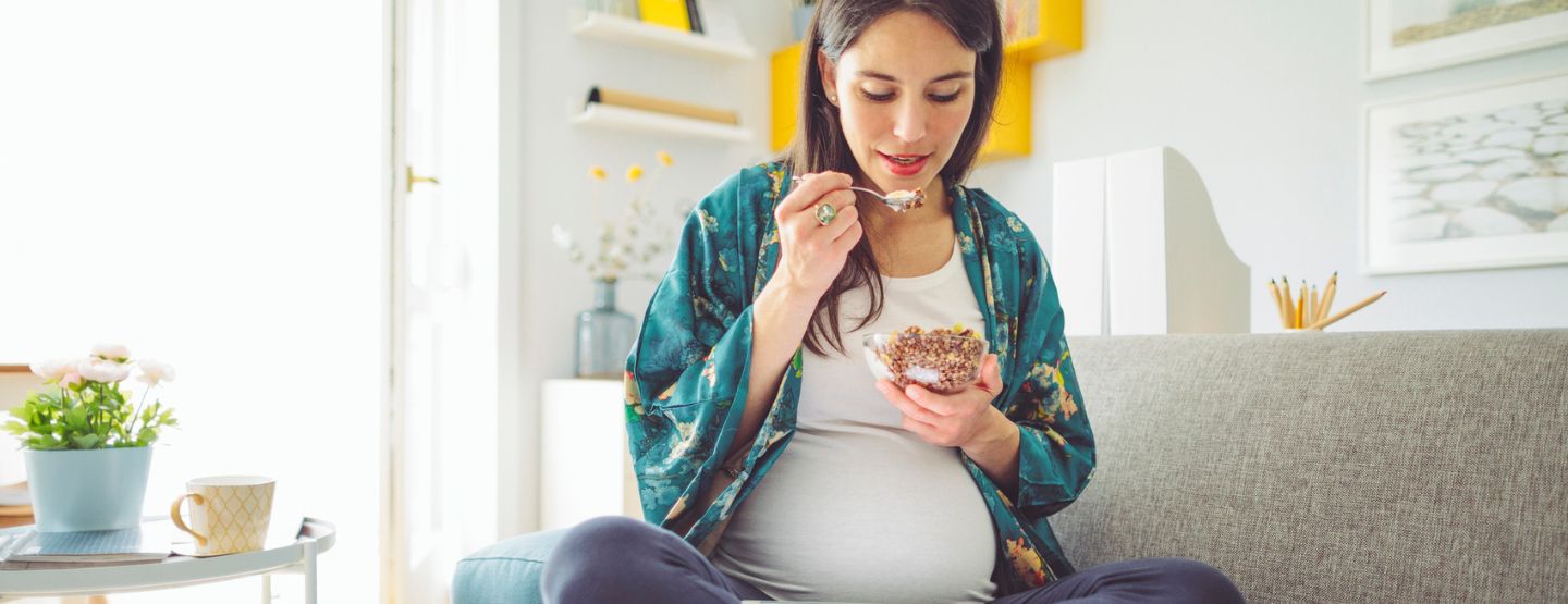Pregnant woman eating bowl of food while sitting on couch.
