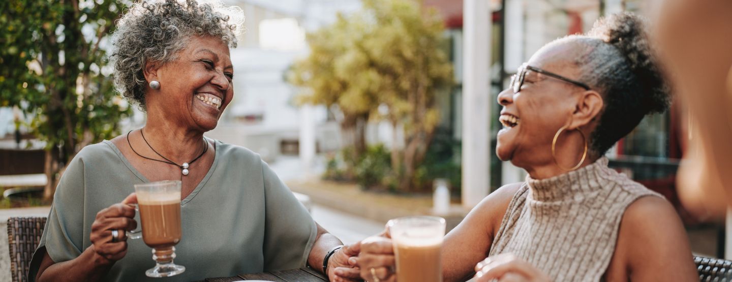 Two mature women laughing while drinking coffee on outdoor patio. 