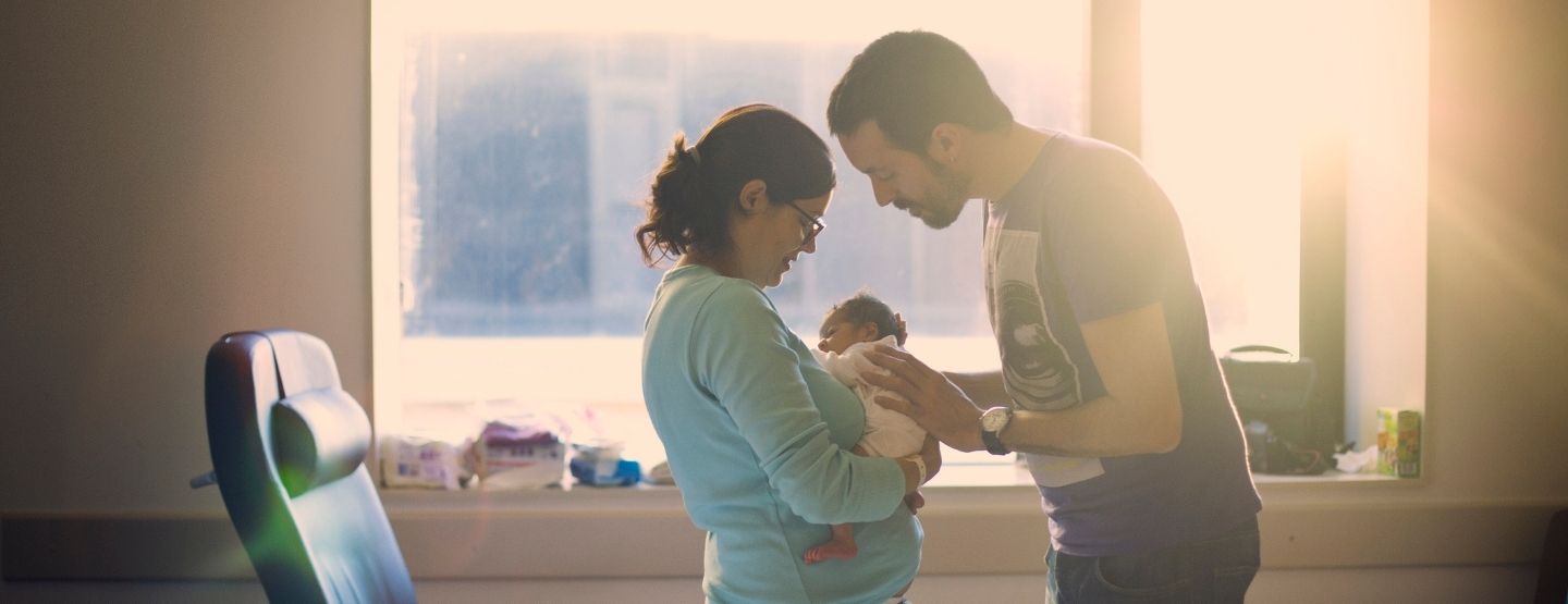 Parents look at at their new baby in the hospital room as sun comes in through the window.