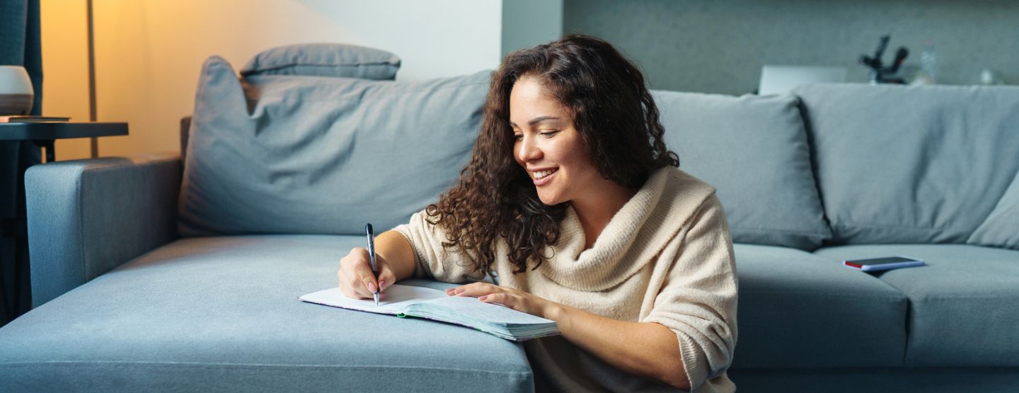 Woman sitting by the couch writing in a notebook.