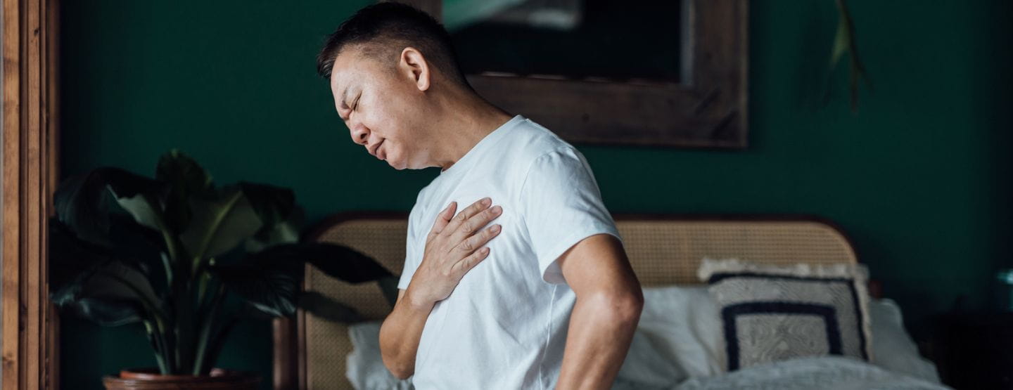 Senior Asian man with eyes closed holding his chest in discomfort, suffering from chest pain while sitting on bed at home. 