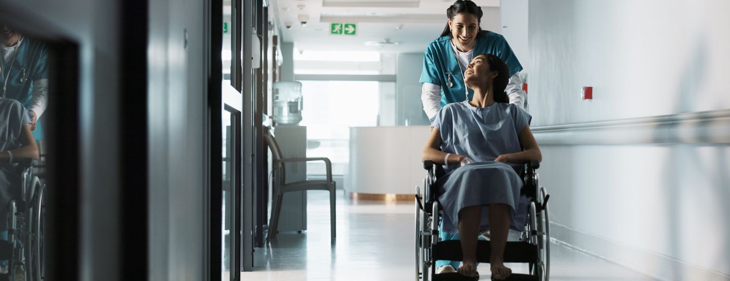 Patient being wheeled down hospital hallway in wheelchair by nurse. 