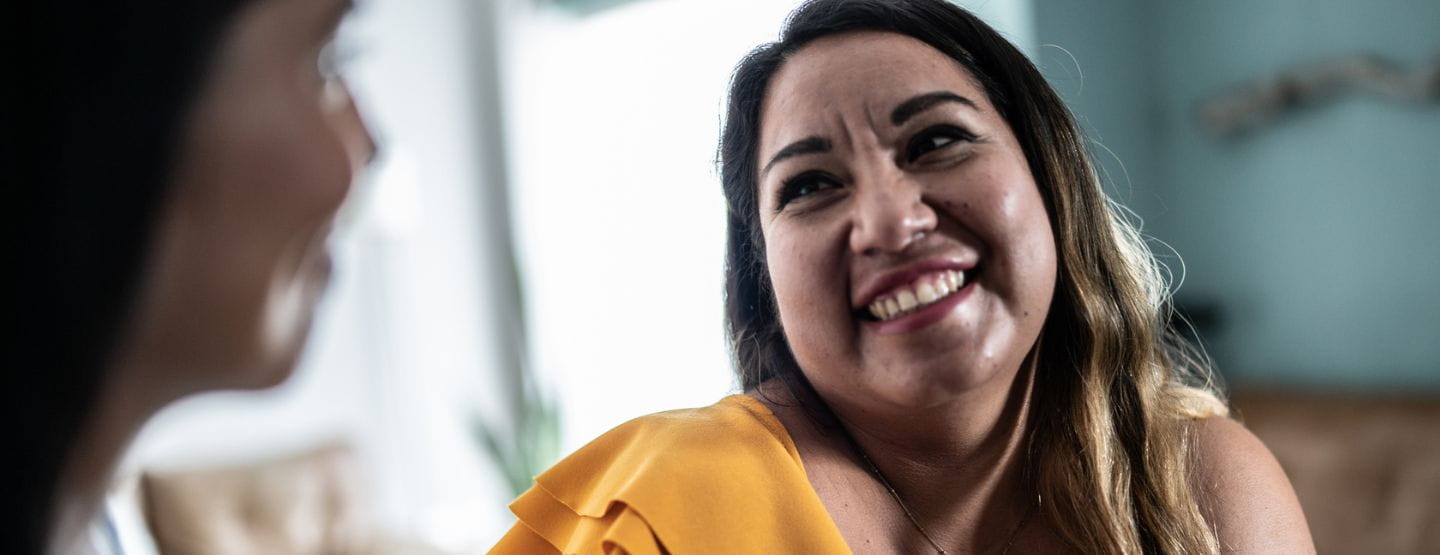 Close up of woman with dark hair and a yellow top smiling to a blurred person slightly off camera
