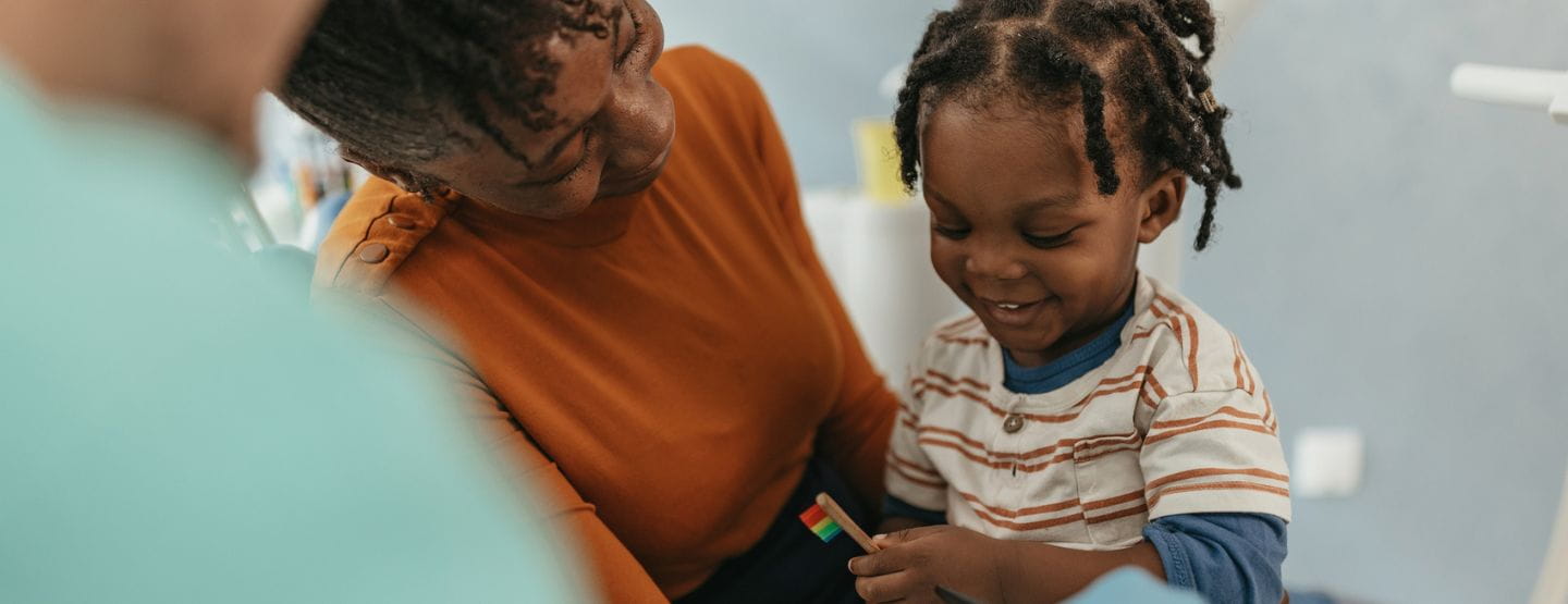 Young child smiles holding new toothbrush and sitting on moms lap at the dentist's office.