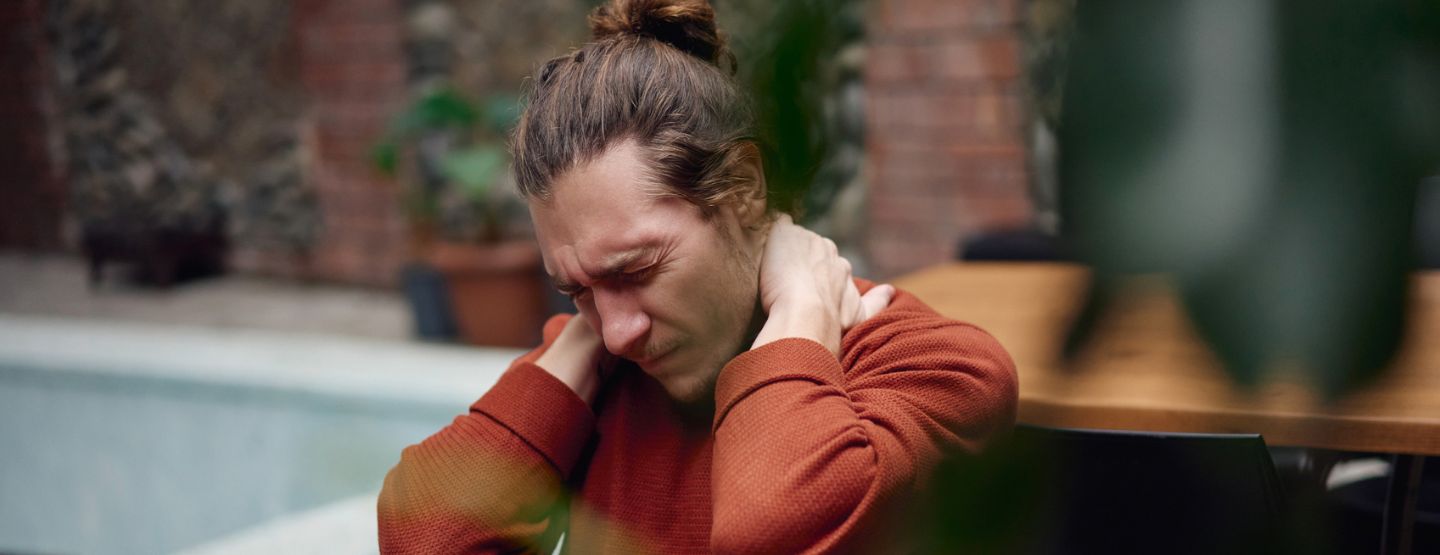 Man winces in pain holding the back of his neck while sitting at a table. 