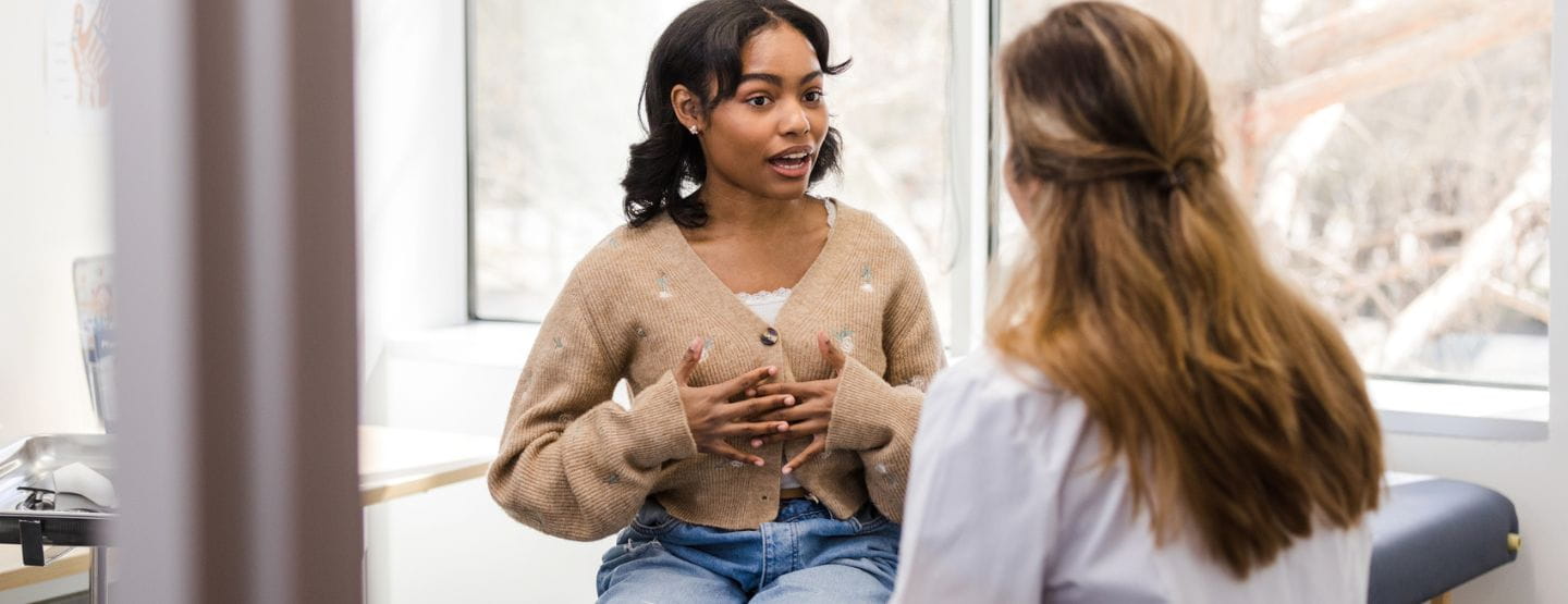 Young adult woman talks to provider while sitting on exam table. 