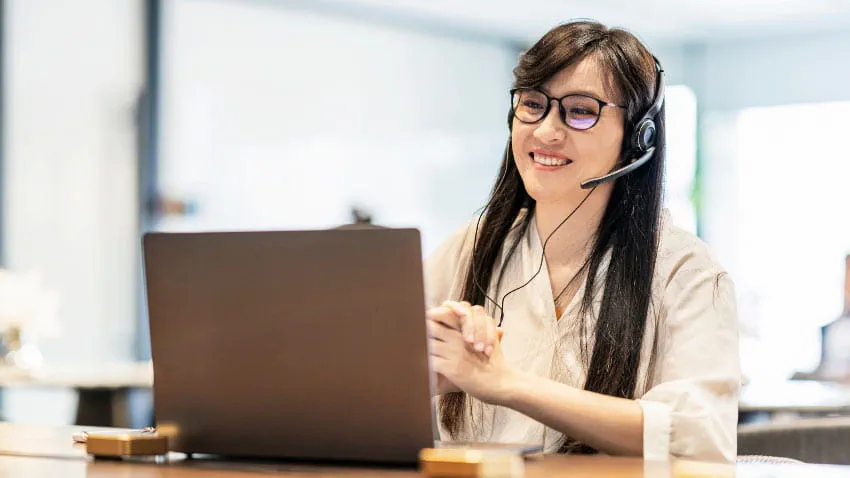 A young woman smiling while wearing a headset and looking at a laptop.