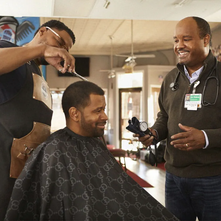 Dr. Brian Dolsey holds a blood pressure cuff next to a man who is getting his hair trimmed by a barber during a community health event