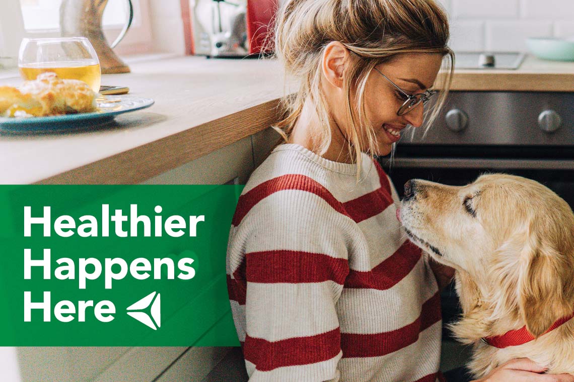 A young woman sits with her dog in the kitchen. Overlay text reads "Healthier Happens Here."