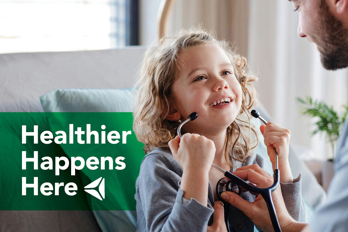 A school-aged girl plays with a doctor's stethoscope. Overlay text reads "Healthier Happens Here."