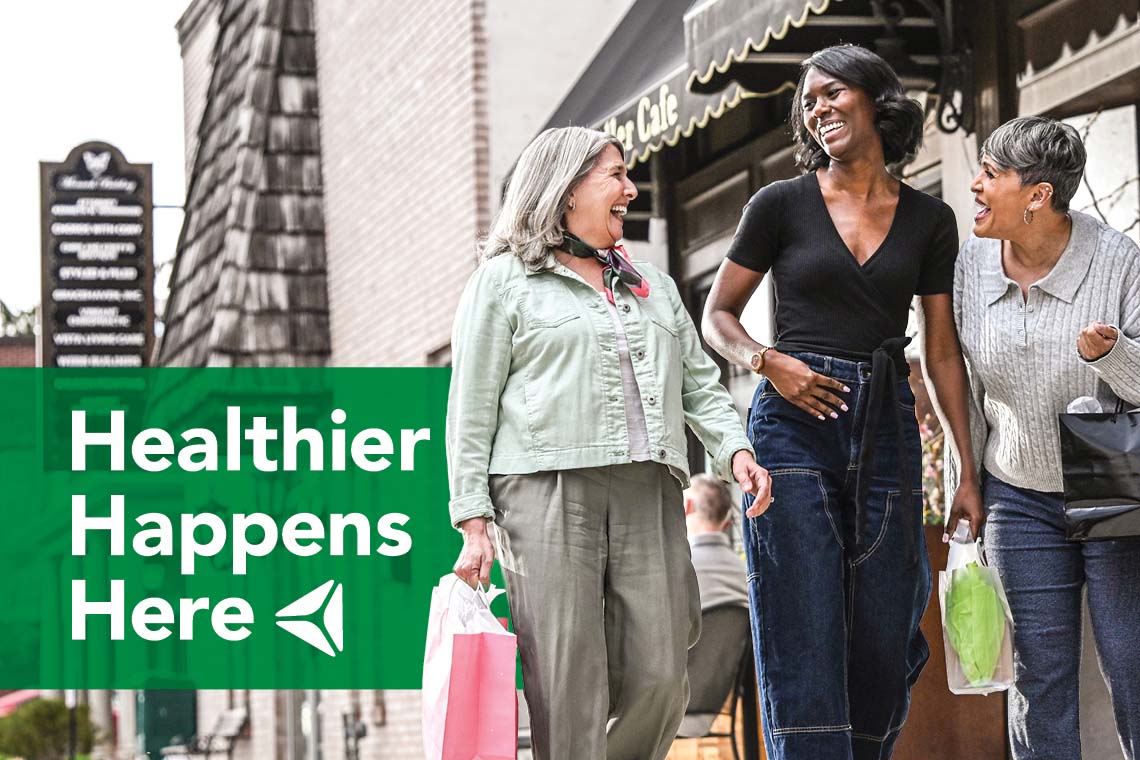 A trio of women hold shopping bags outside of a boutiqe. Overlay text reads "Healthier Happens Here."