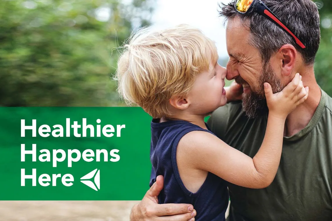 A father embraces his young son. Overlay text reads "Healthier Happens Here."
