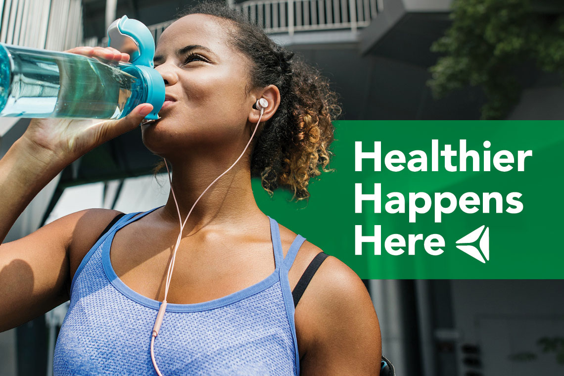 A young woman drinks from her water bottle while exercising. Overlay text reads "Healthier Happens Here."