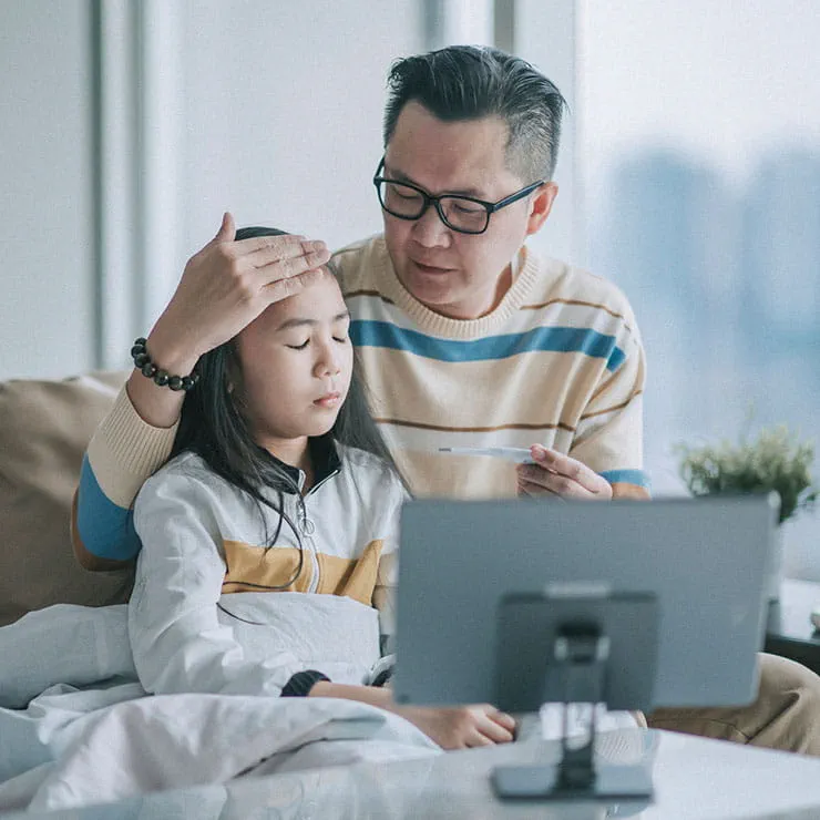 a father feels his daughter's forehead for fever during a virtual urgent care appointment on a computer tablet