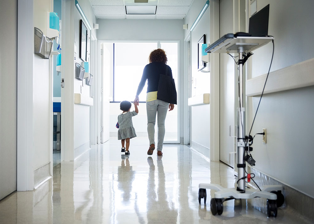 A child and healthcare provider walk hand in hand down a hospital corridor