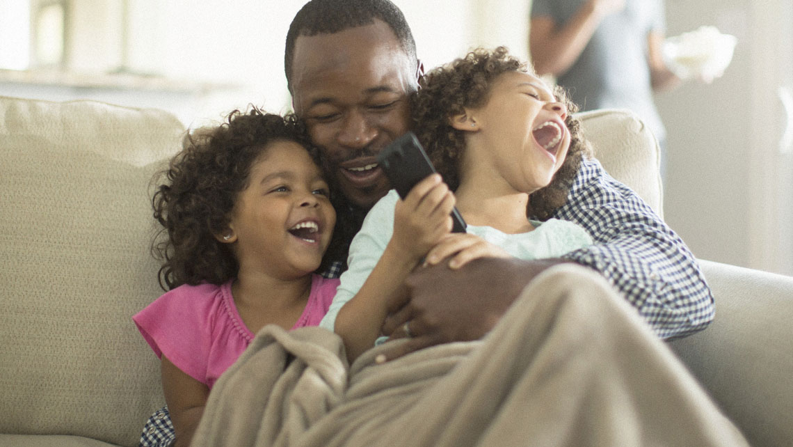 a grandfather hugs his two young granddaughters at home on the couch while watching television together