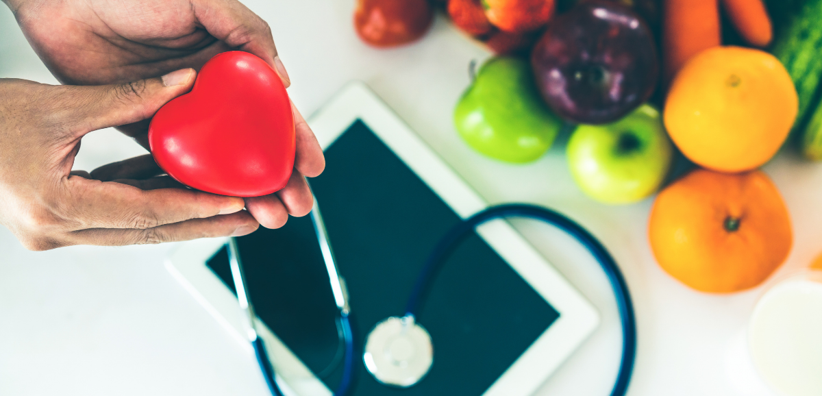 A persons hands holding a red heart above a stethoscope, a tablet, and other fruits and vegetables.