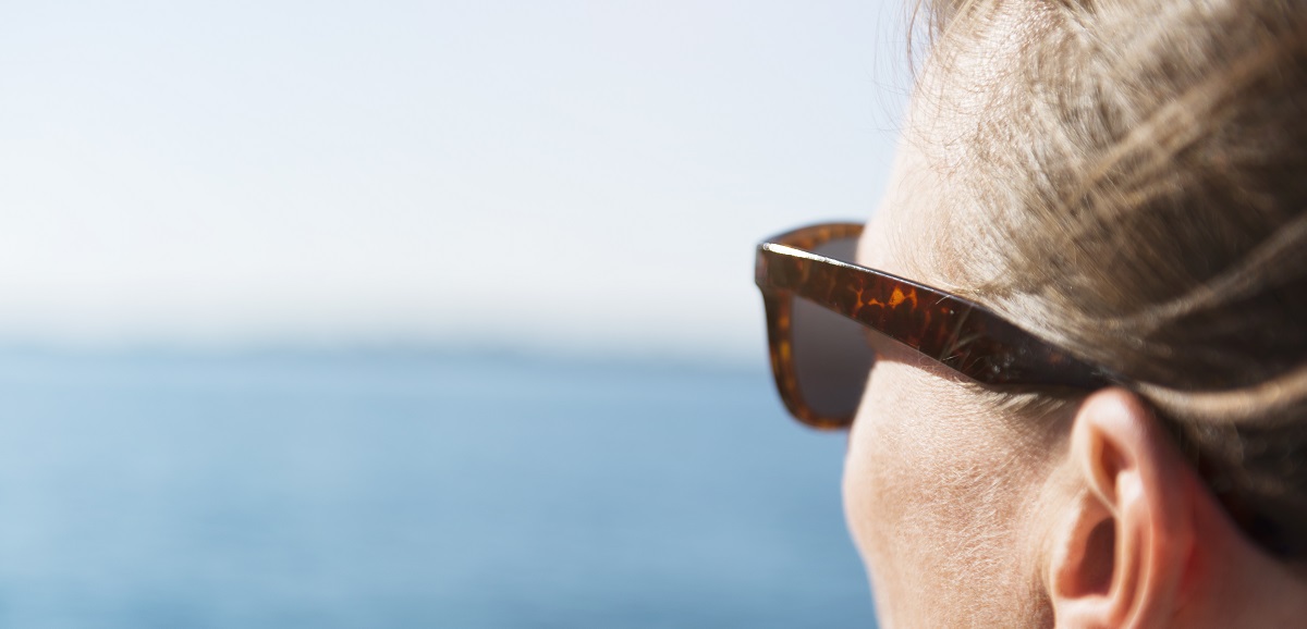 A close up profile image of a woman wearing sunglasses while staring out at the ocean.